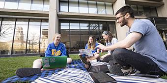 four engineering students sit on blankets on the lawn outside their building to study together with laptops. 
